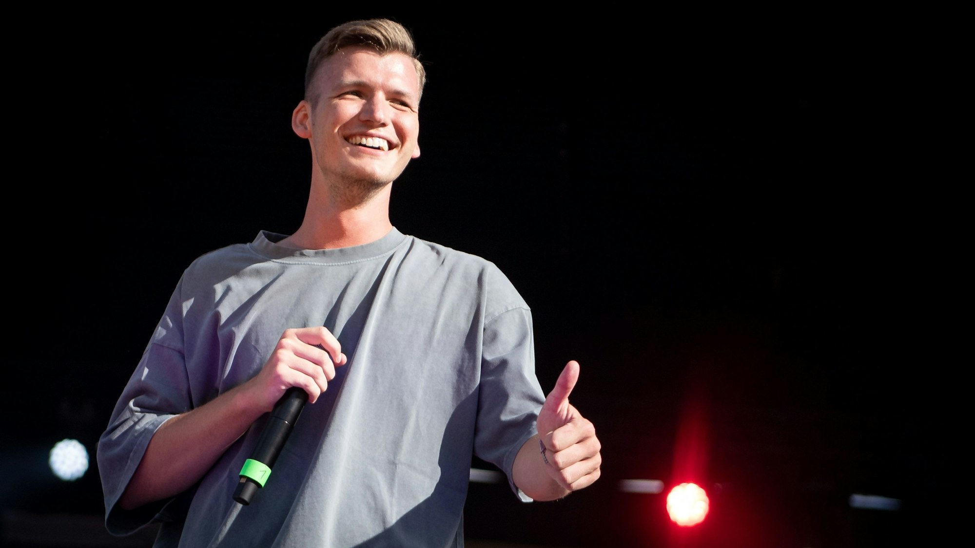 Ein junger Mann im T-Shirt hat ein Mikrofon inder Hand, lächelt breit und hebt den Daumen.