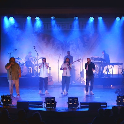 Eine Band mit vier Sängern im Vordergrund und mehreren Instrumentalisten steht auf der blau illuminierten Bühne des Euskirchener Theaters.