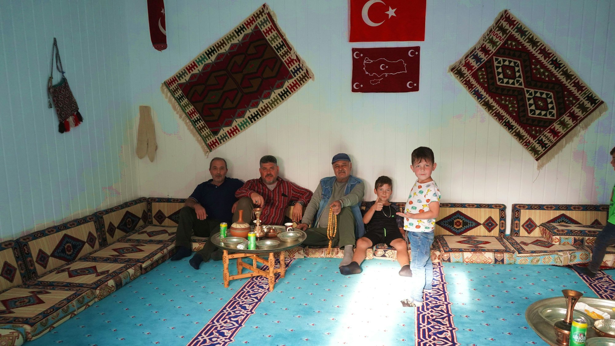 Mehrere Männer und ein Junge sitzen am Rande des Gebetsraums in der Moschee der Türkisch-Islamischen Gemeinde Euskirchen. Ein zweiter Junge steht bei ihnen. An der Wand hängen Teppiche und eine türkische Flagge.
