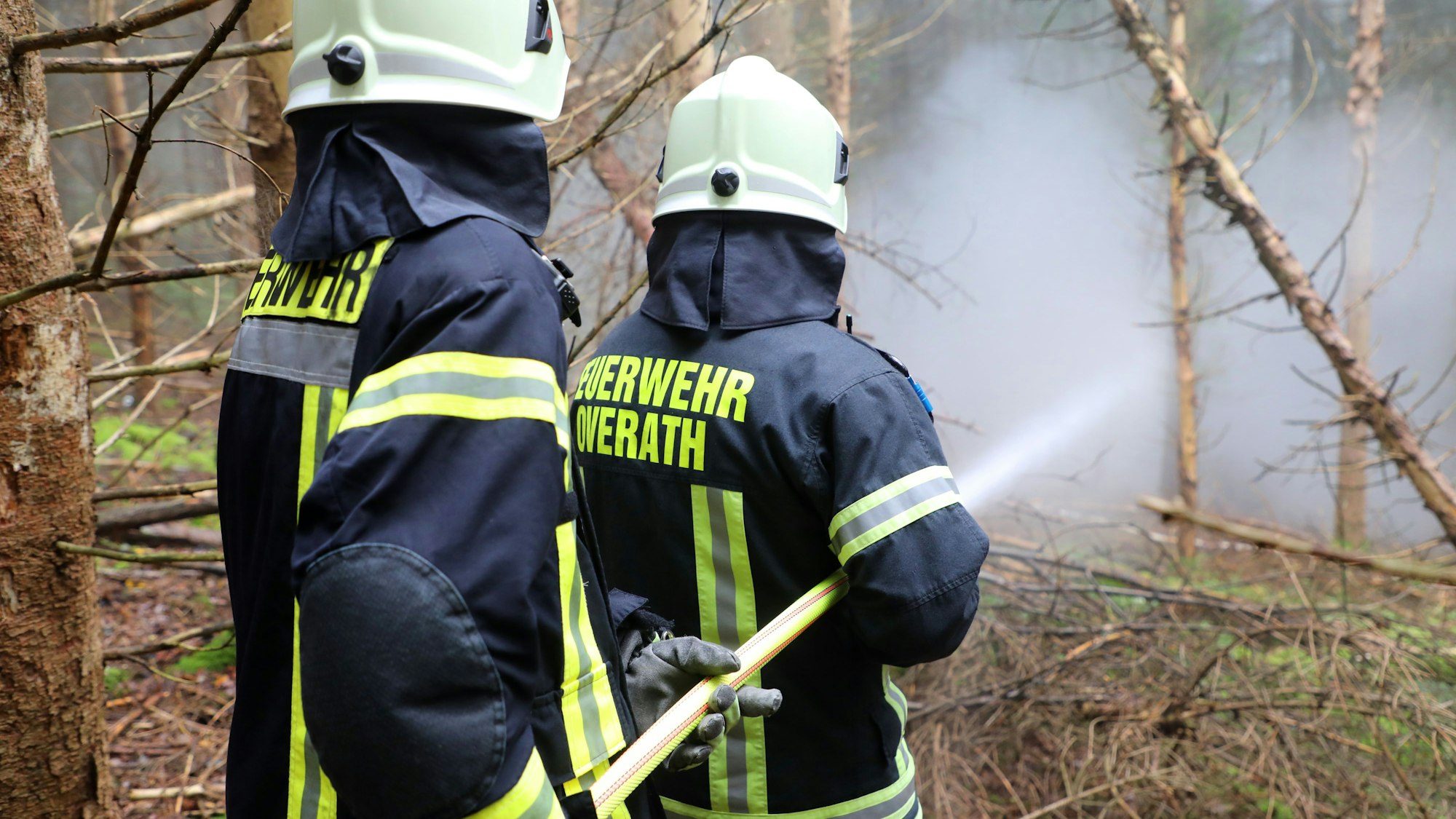 Zwei Feuerwehrleute der Feuerwehr Overath halten einen C-Schlauch auf Übungsrauch in einem Fichtenwald.