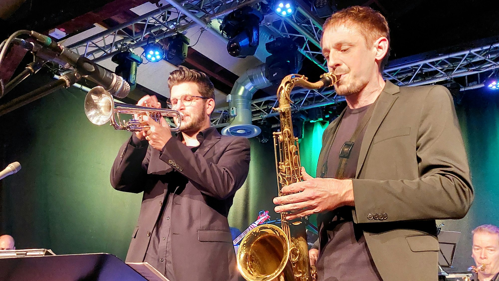 Ein junger Mann mit Trompete und ein weiterer mit Saxofon stehen im Rampenlicht auf einer Bühne, beide tragen Anzüge.