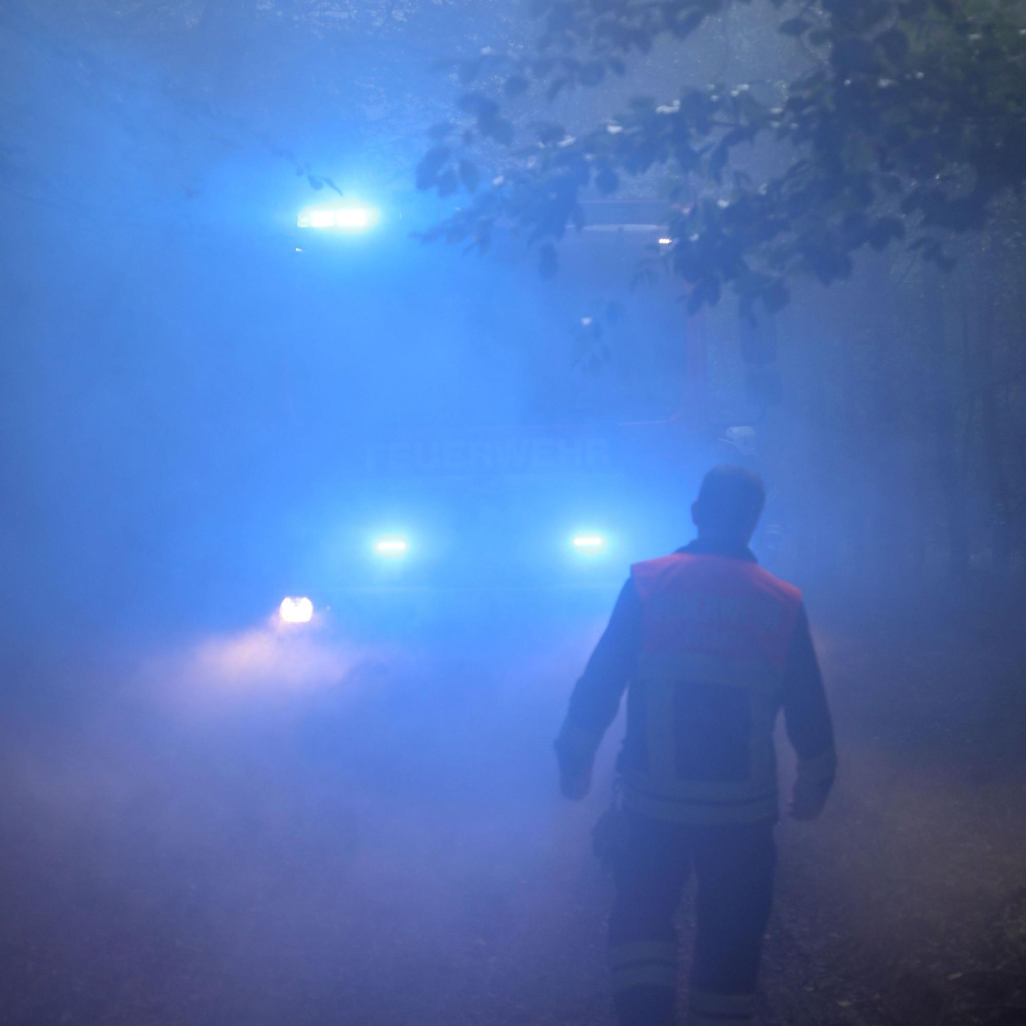 Ein Feuerwehrmann geht durch Rauch auf ein Feuerwehrfahrzeug mit Blaulicht zu.