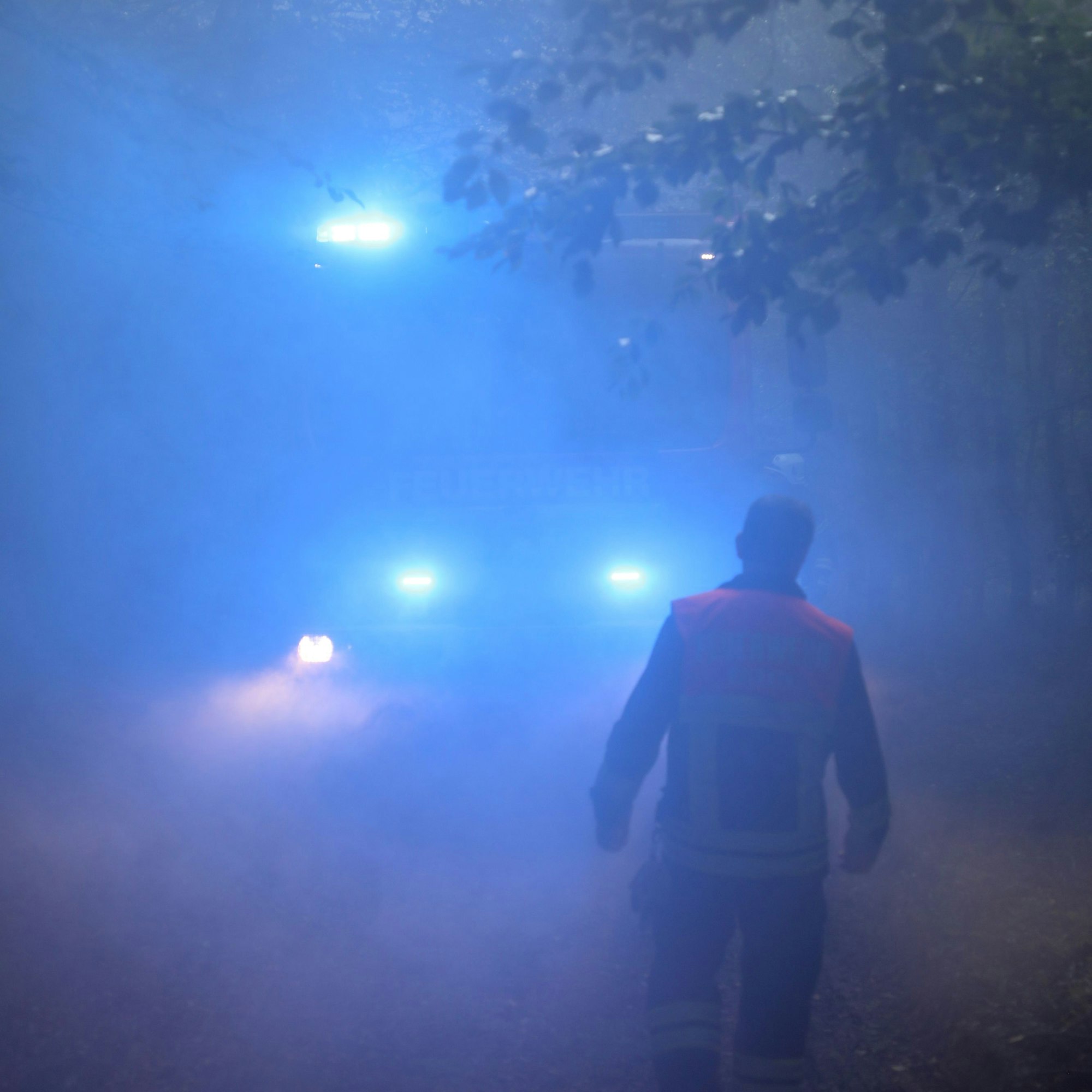 Ein Feuerwehrmann geht durch Rauch auf ein Feuerwehrfahrzeug mit Blaulicht zu.