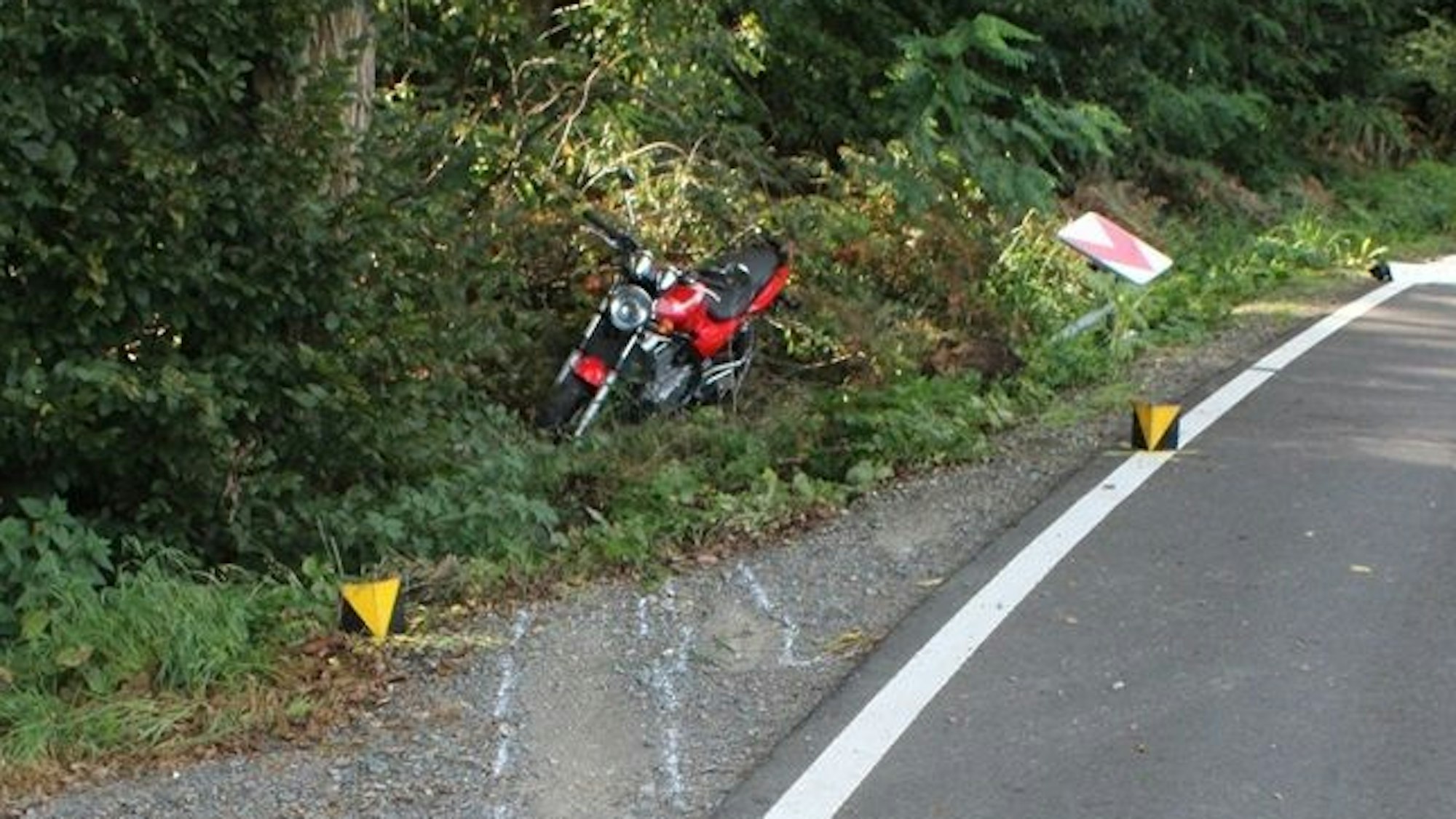 Ein Motorrad steht nach einem Unfall im Straßengraben, Markierungen der Polizei sind auf der Straße zu sehen und ein herausgerissenes Verkehrsschild.