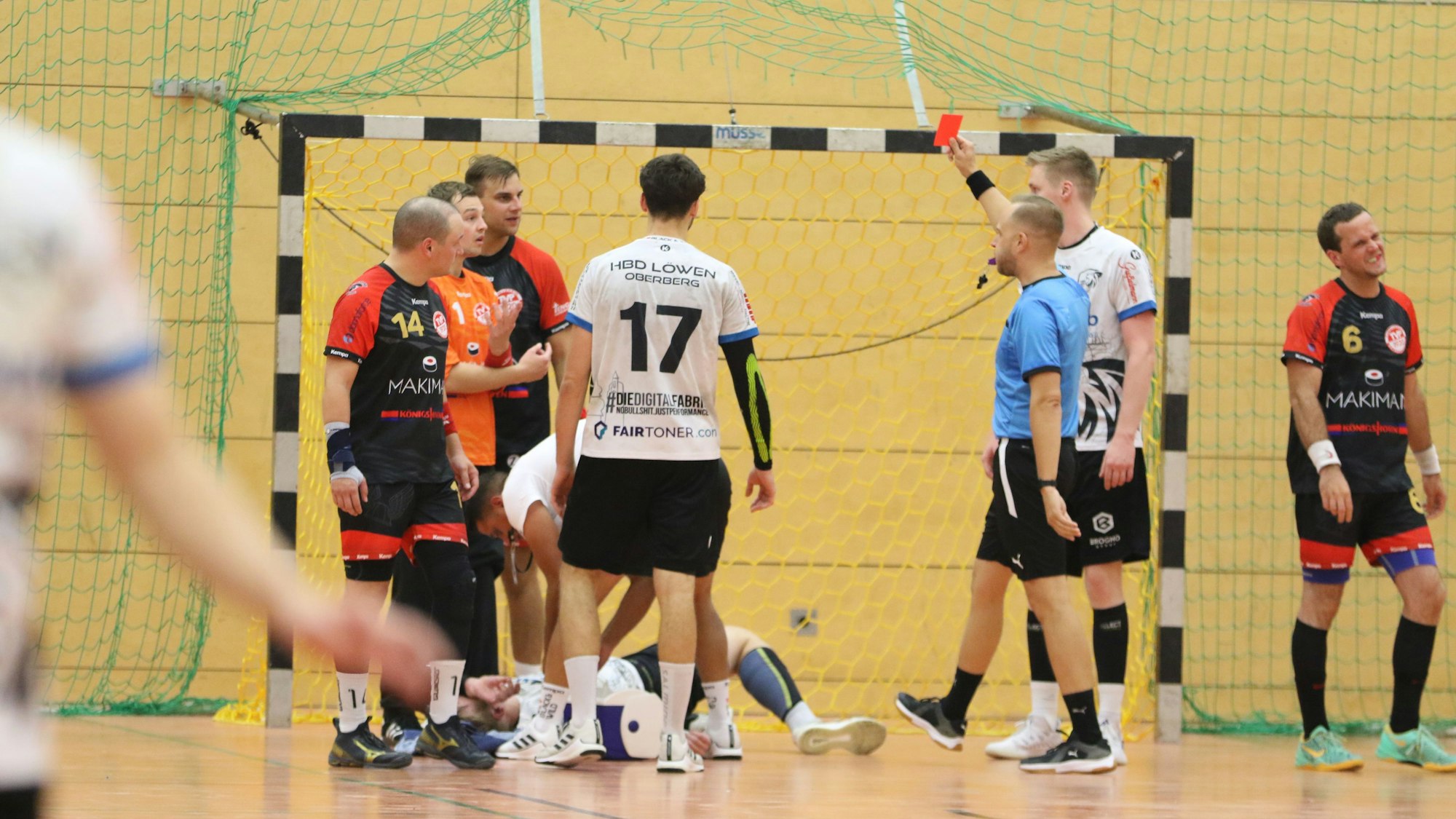 Ein Schiedsrichter zeigt einem Handballspieler die Rote Karte. Die Mitspieler sind fassungslos. Ein gegnerischer Spieler wird behandelt.
