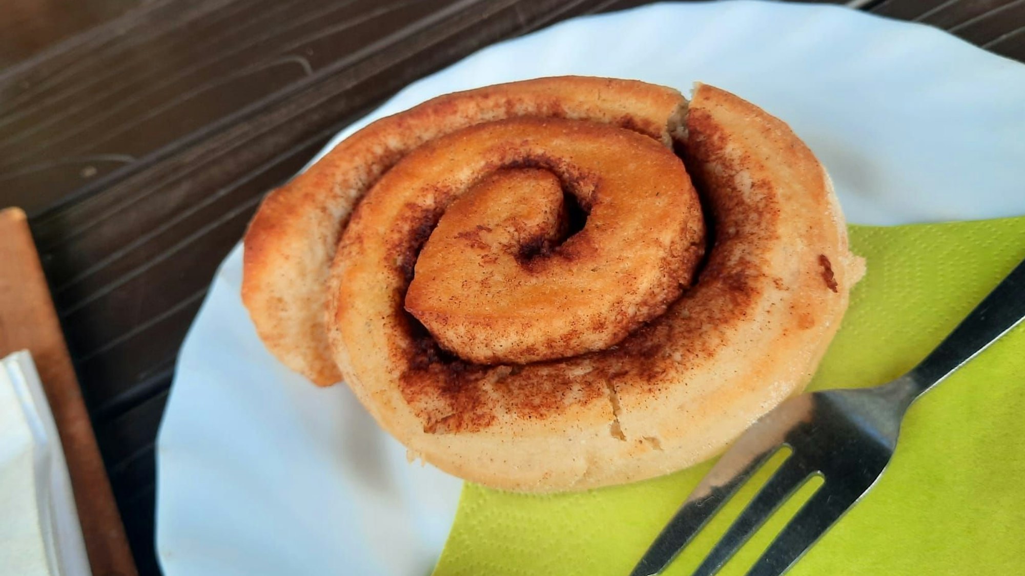 Die Zimtschnecke ist auf einem weißen Teller serviert, daneben eine grüne Serviette mit Kuchengabel.