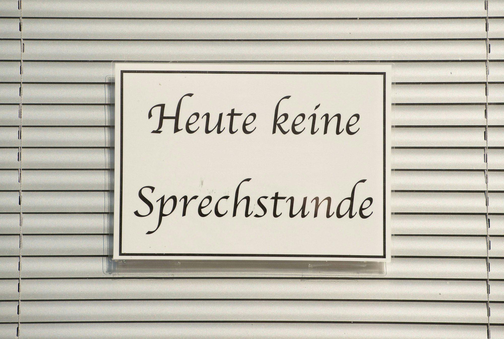 Ein Schild mit der Aufschrift ‚Heute keine Sprechstunde‘ hängt im Fenster einer Arztpraxis vor einer Jalousie.
