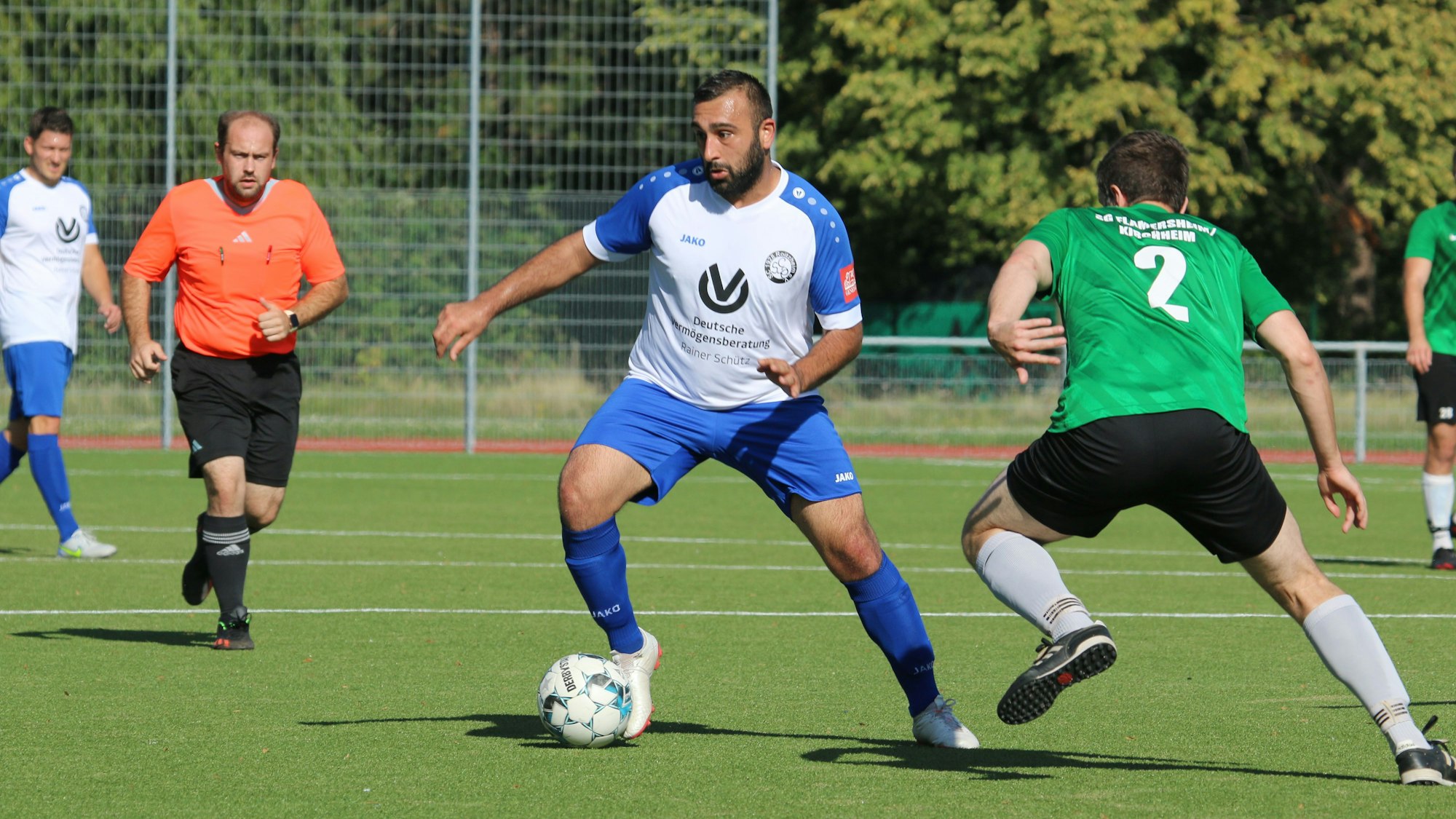 Ein Fußballer führt den Ball, ein Gegner hat ihn im Visier. Der Schiedsrichter beobachtet die Szene.