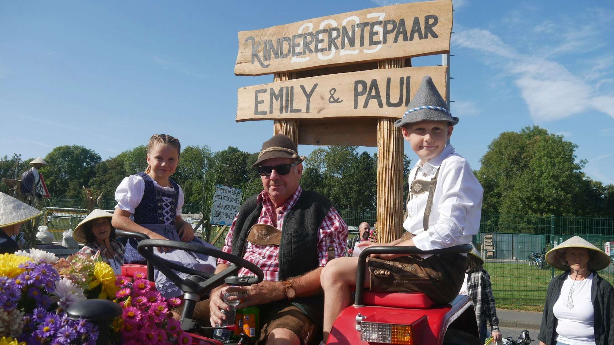 Auf einem roten, blumengeschmückten Trecker sitzt ein älterer Mann mit Lederhose, Weste und rotkariertem Hemd. Auf den Sitzen neben ihm ein Junge und ein Mädchen, ebenfalls in bayrischer Tracht.