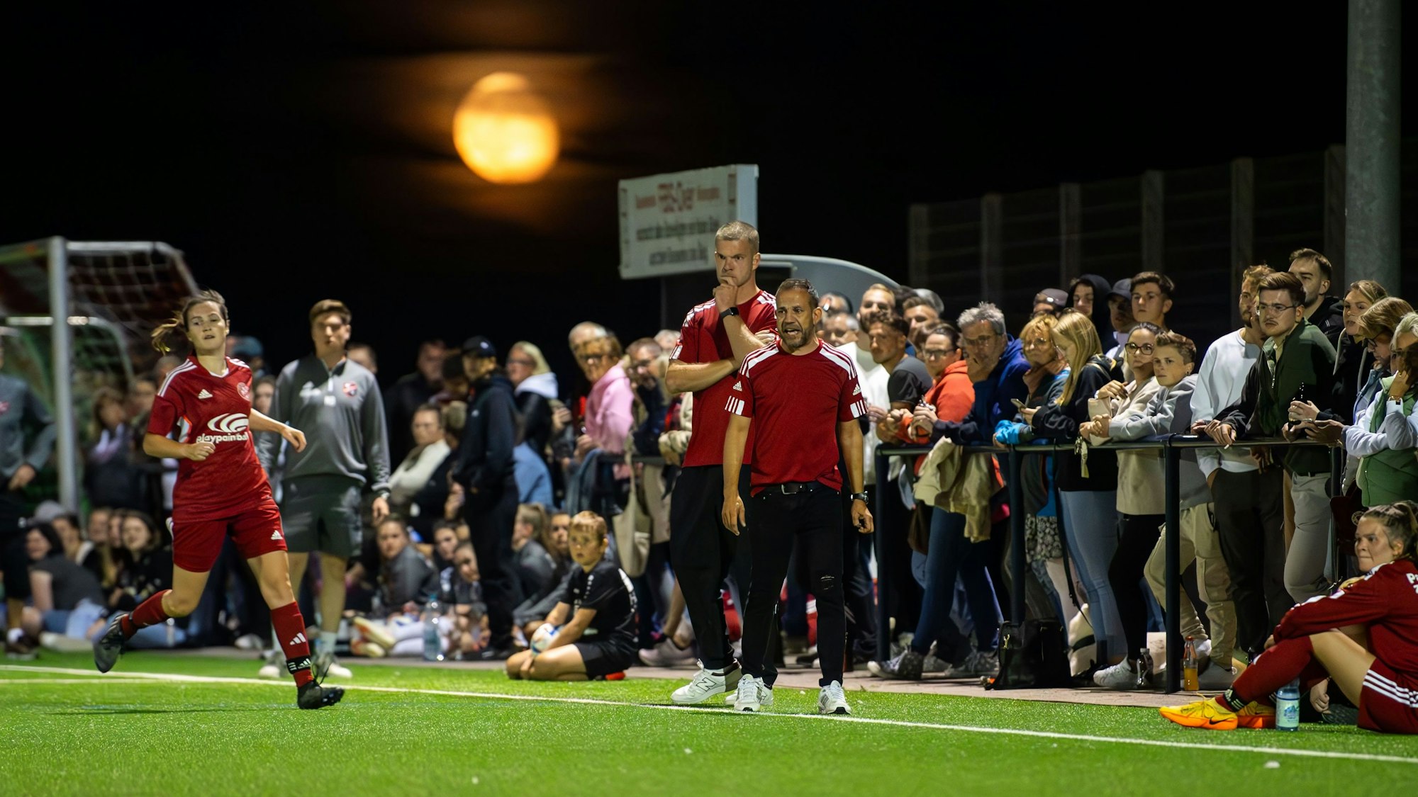 Zuschauer stehen direkt hinter dem Zülpicher Trainerduo am Spielfeldrand. Im Hintergrund der blutrote Mond.
