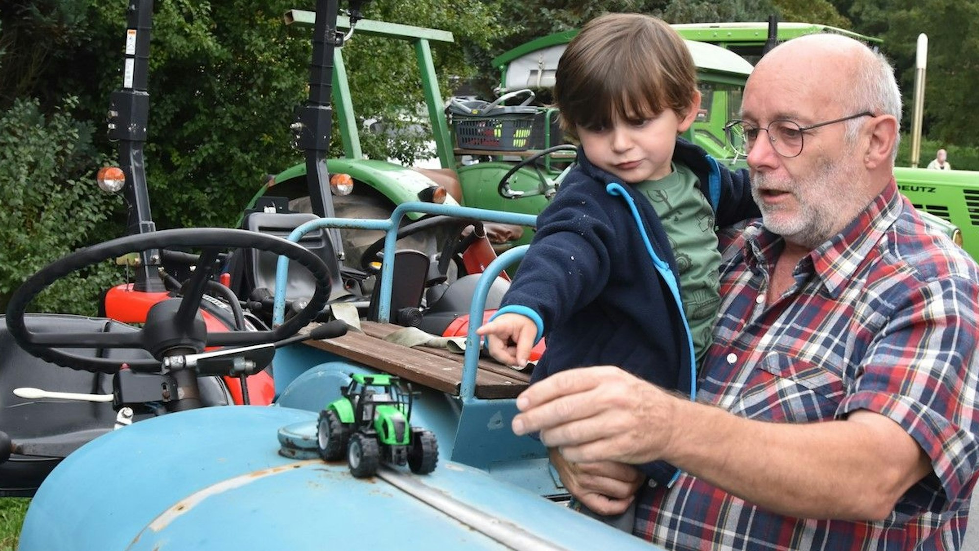 Ein kleiner Junge auf dem Arm seines Großvaters zeiigt auf einen kleinen Spielzeugtraktor, der auf der Motorhaube eines richtigen Traktors steht.