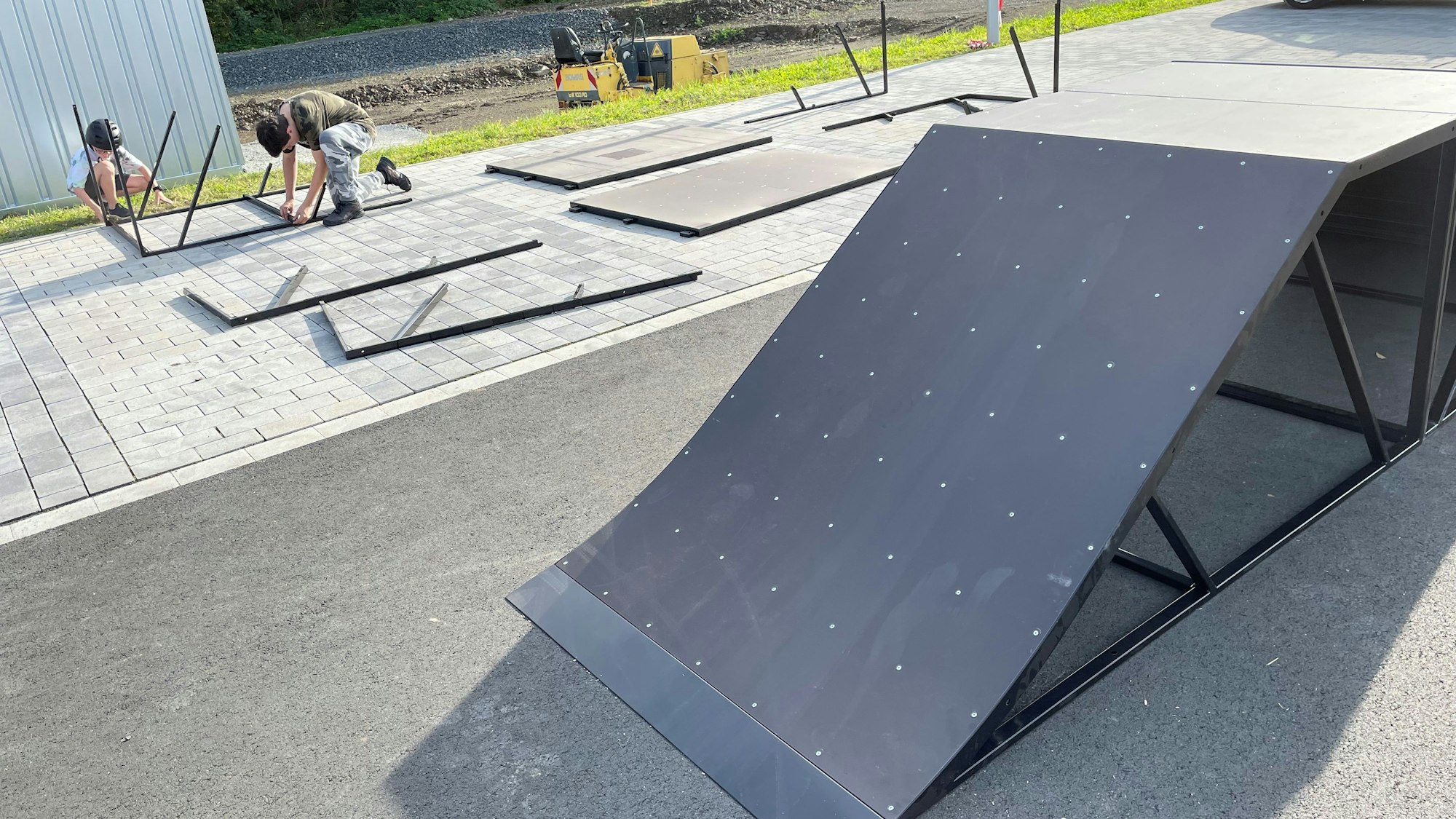 Die Rampe eines mobilen Skateparks ist zu sehen. Im Hintergrund schrauben zwei Jugendliche weitere Elemente zusammen.