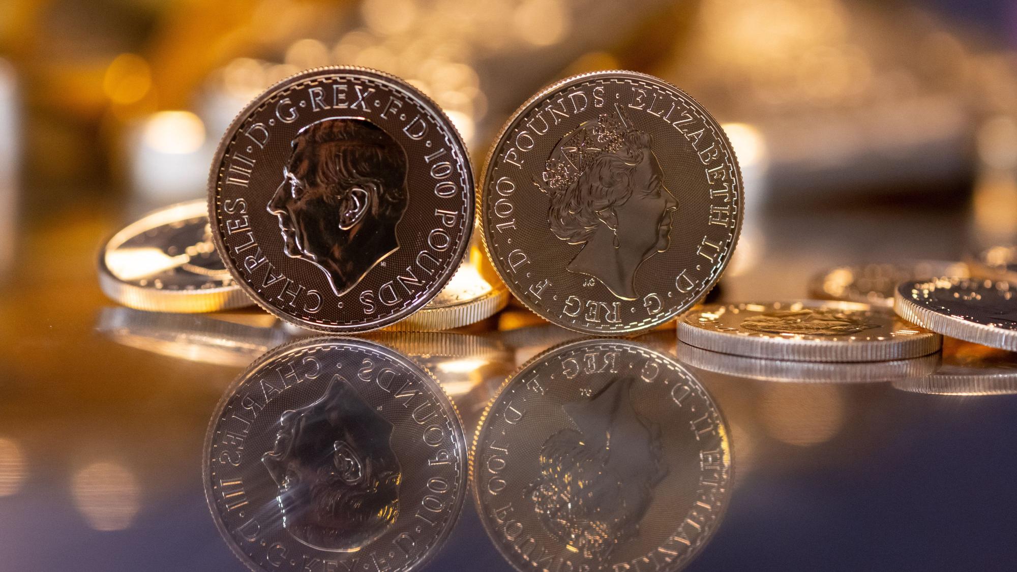 Goldbarren und Goldmünzen in unterschiedlicher Größe liegen beim Edelmetallhändler Pro Aurum in einem Tresor auf einem Tisch. Im Vordergrund ist die Goldmünze "Britannia" zu sehen (links mit dem Motiv König Charles III., rechts mit dem Motiv Königin Elizabeth II.)