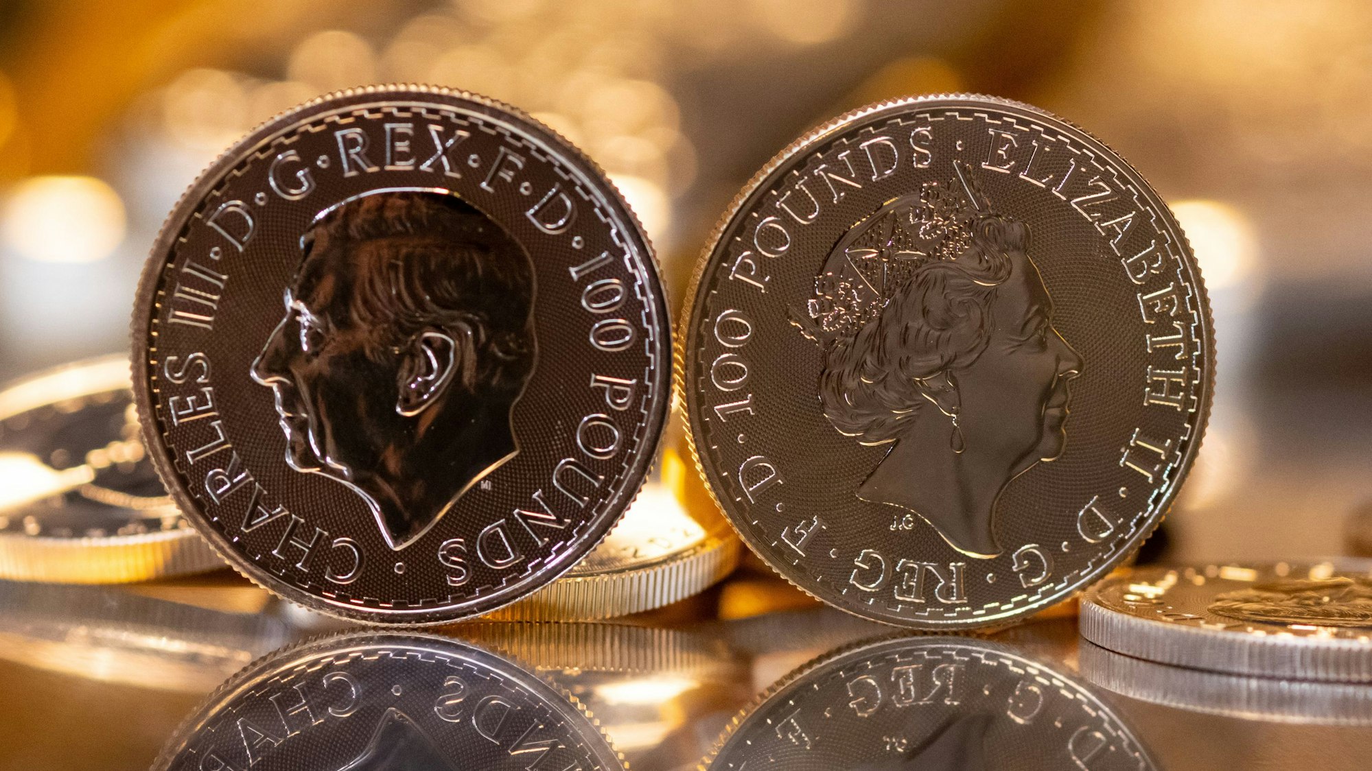 Goldbarren und Goldmünzen in unterschiedlicher Größe liegen beim Edelmetallhändler Pro Aurum in einem Tresor auf einem Tisch. Im Vordergrund ist die Goldmünze "Britannia" zu sehen (links mit dem Motiv König Charles III., rechts mit dem Motiv Königin Elizabeth II.)
