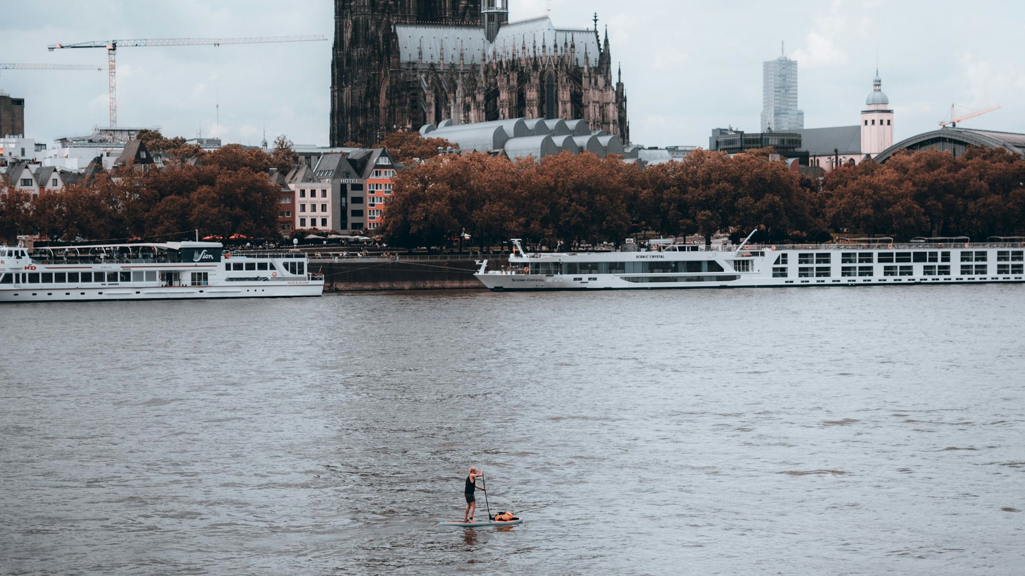 Ein Stand-Up-Paddler ist am Abend auf dem Rhein vor dem Rheinboulevard unterwegs, im Hintergrund ist der Dom zu sehen.