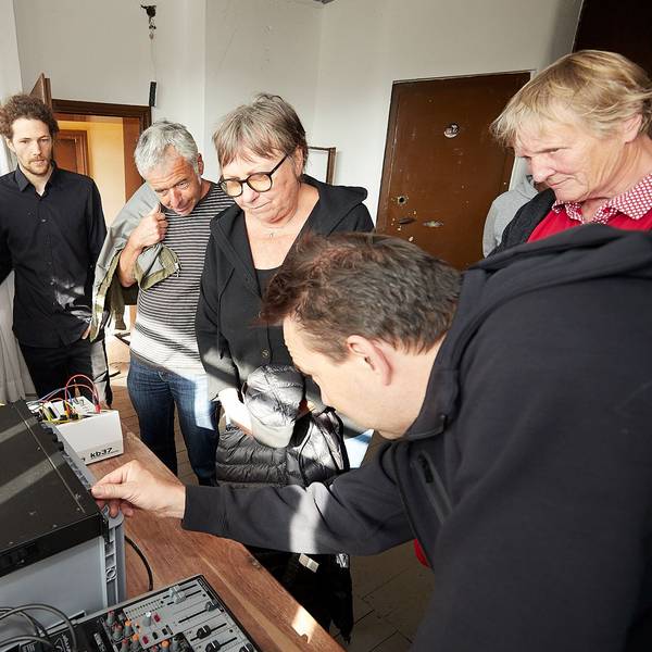 Eine Gruppe Menschen steht um einen Tisch, auf dem elektronische Geräte aufgebaut sind. Ein Mann bedient einen Regler.