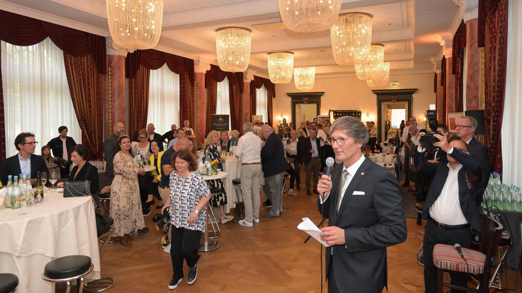 Jörg Stricker steht im Ballsaal vor vielen Menschen und hält ein Mikrofon.