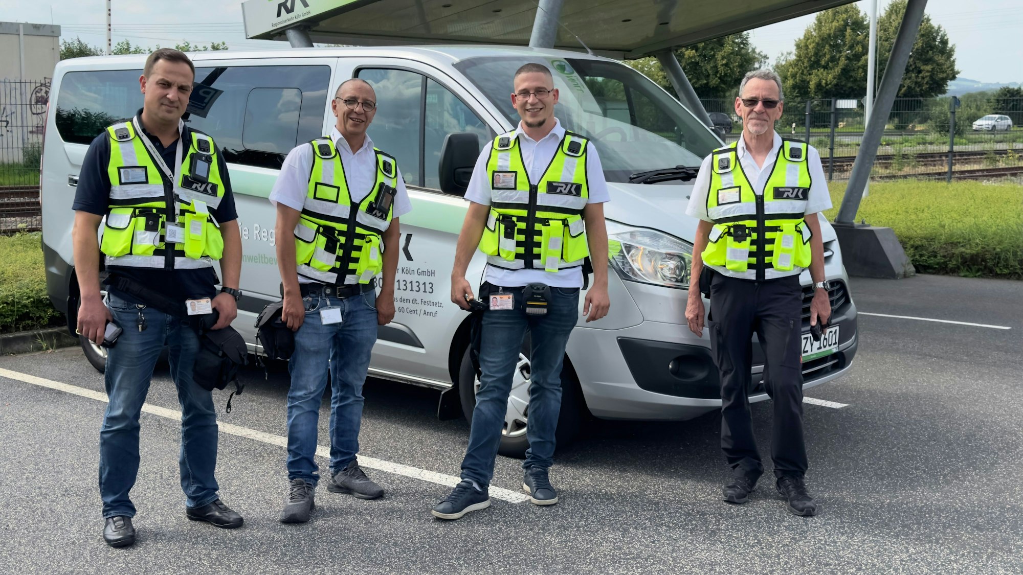 Vier Service-Mitarbeiter der Regionalverkehr Köln GmbH stehen mit gelben Westen vor einem Einsatzfahrzeug. Zwei der Kontrolleure tragen Body-Cams an ihren Westen.