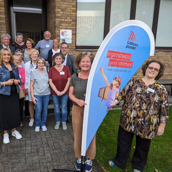Eine Gruppe von Frauen und Männern vor einem Haus, im Fordergrund zwei Frauen an einer Flagge mit der Aufschrift Lotsenpunkt.