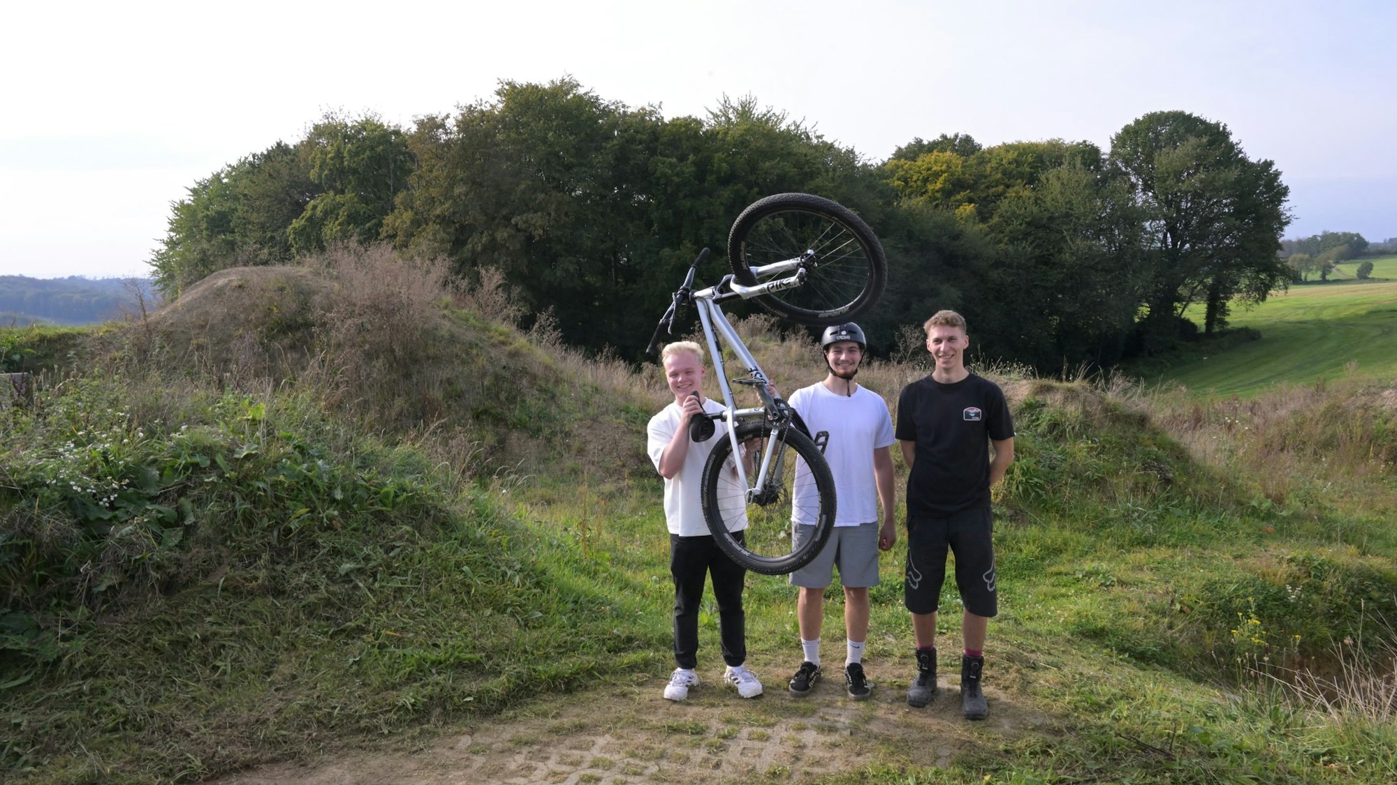 Vor der gerade entstehenden Dirtanlage stehen drei junge Sportler.
