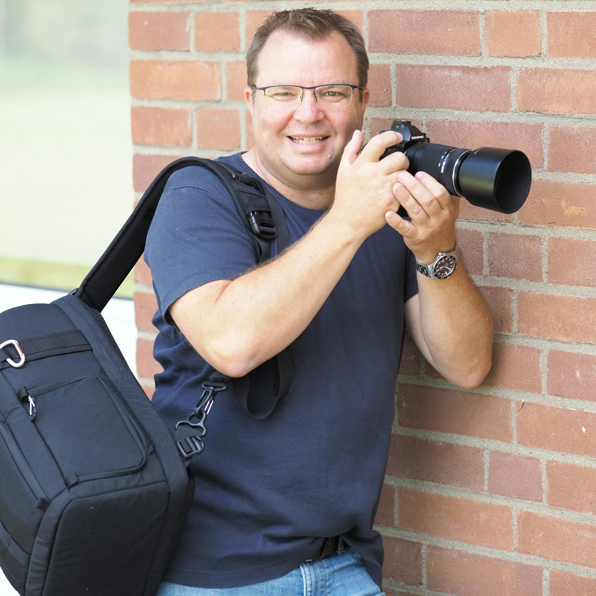 Ein Mann mit Kamera lehnt an einer Ziegelsteinwand.