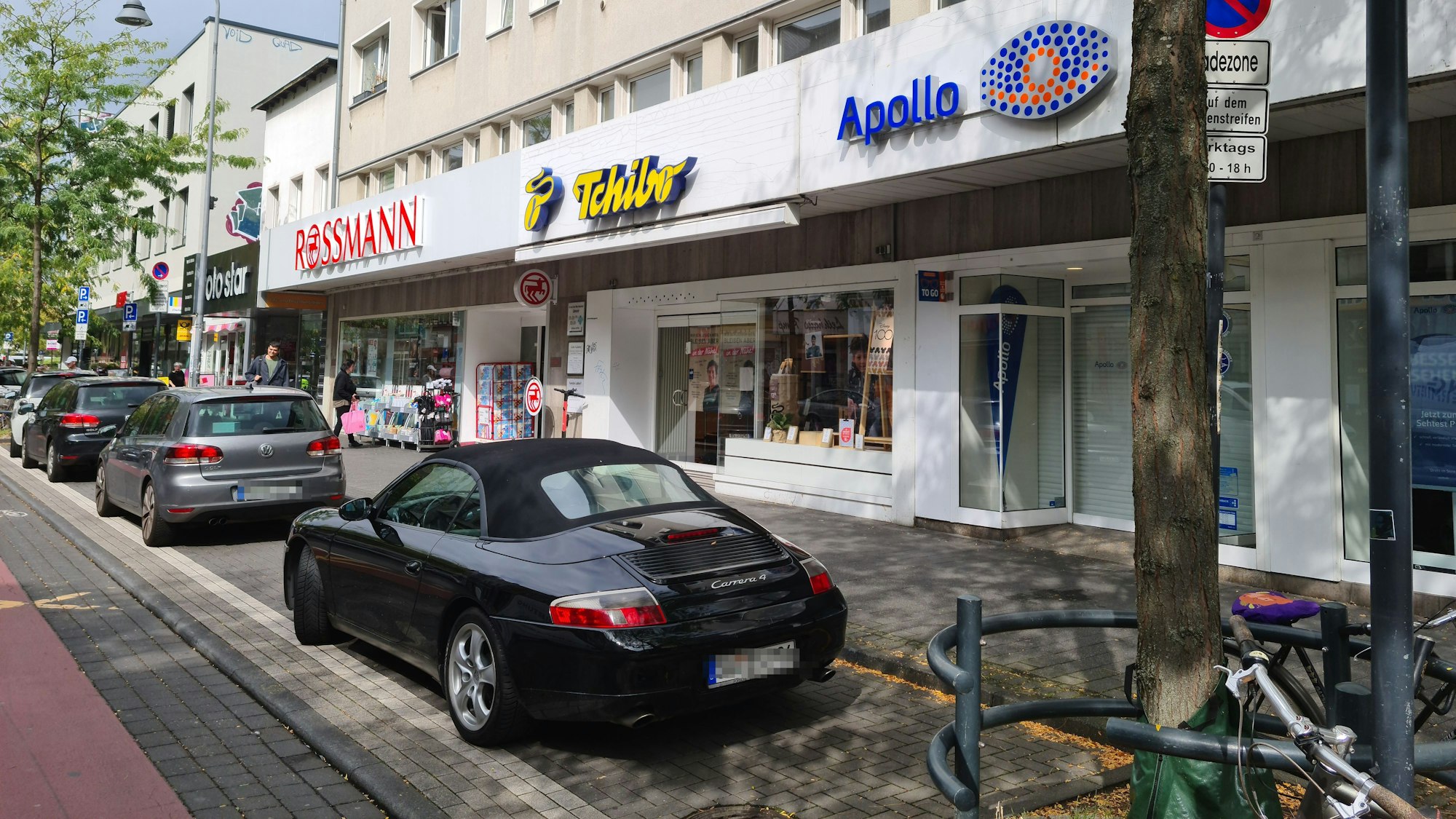 An einer Straße liegen die Filialen von Rossmann, Tchibo und Apollo direkt nebeneinander, vor den Ladenlokalen parken Autos auf dem Parkstreifen an einer Straße.