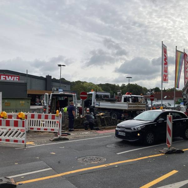 Zu sehen sind Baufahrzeuge und Absperrbaken vor dem Supermarktparkplatz. Daran vorbei fährt ein Kleinwagen.