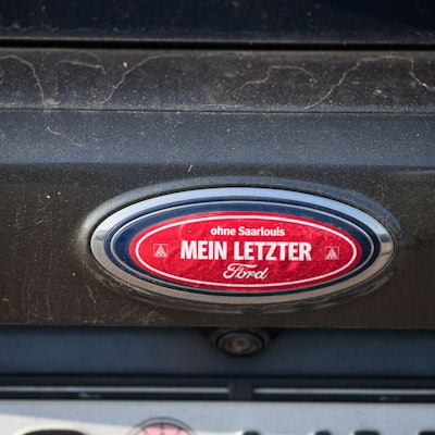 04.10.2023, Saarland, Saarlouis: Ein Aufkleber "ohne Saarlouis mein letzter Ford" klebt auf dem Ford Logo einen Autos, das auf dem dem Parkplatz des Ford Werks in Saarlouis steht. Bei einer Betriebsversammlung hofft die Belegschaft mehr über die Zukunft des Werkes zu erfahren. Foto: Oliver Dietze/dpa +++ dpa-Bildfunk +++