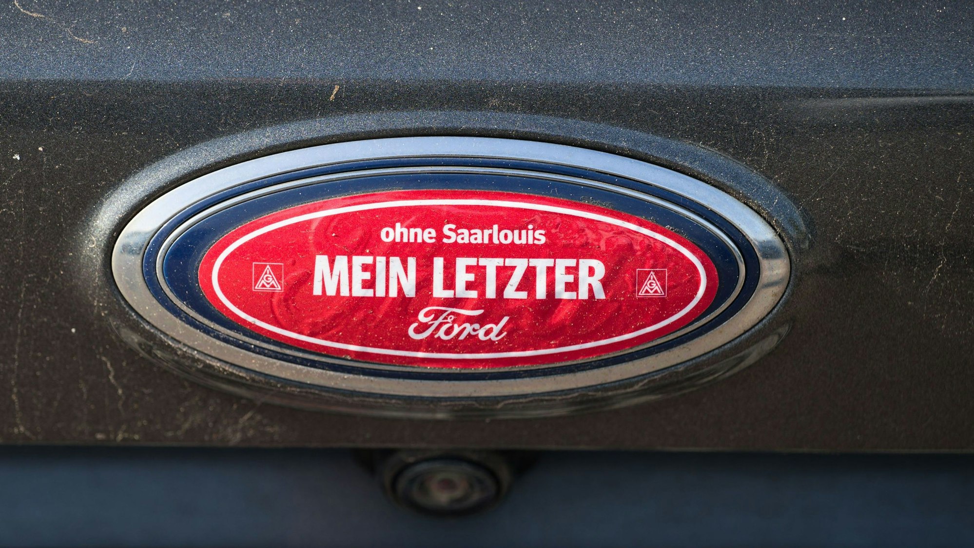 04.10.2023, Saarland, Saarlouis: Ein Aufkleber "ohne Saarlouis mein letzter Ford" klebt auf dem Ford Logo einen Autos, das auf dem dem Parkplatz des Ford Werks in Saarlouis steht. Bei einer Betriebsversammlung hofft die Belegschaft mehr über die Zukunft des Werkes zu erfahren. Foto: Oliver Dietze/dpa +++ dpa-Bildfunk +++