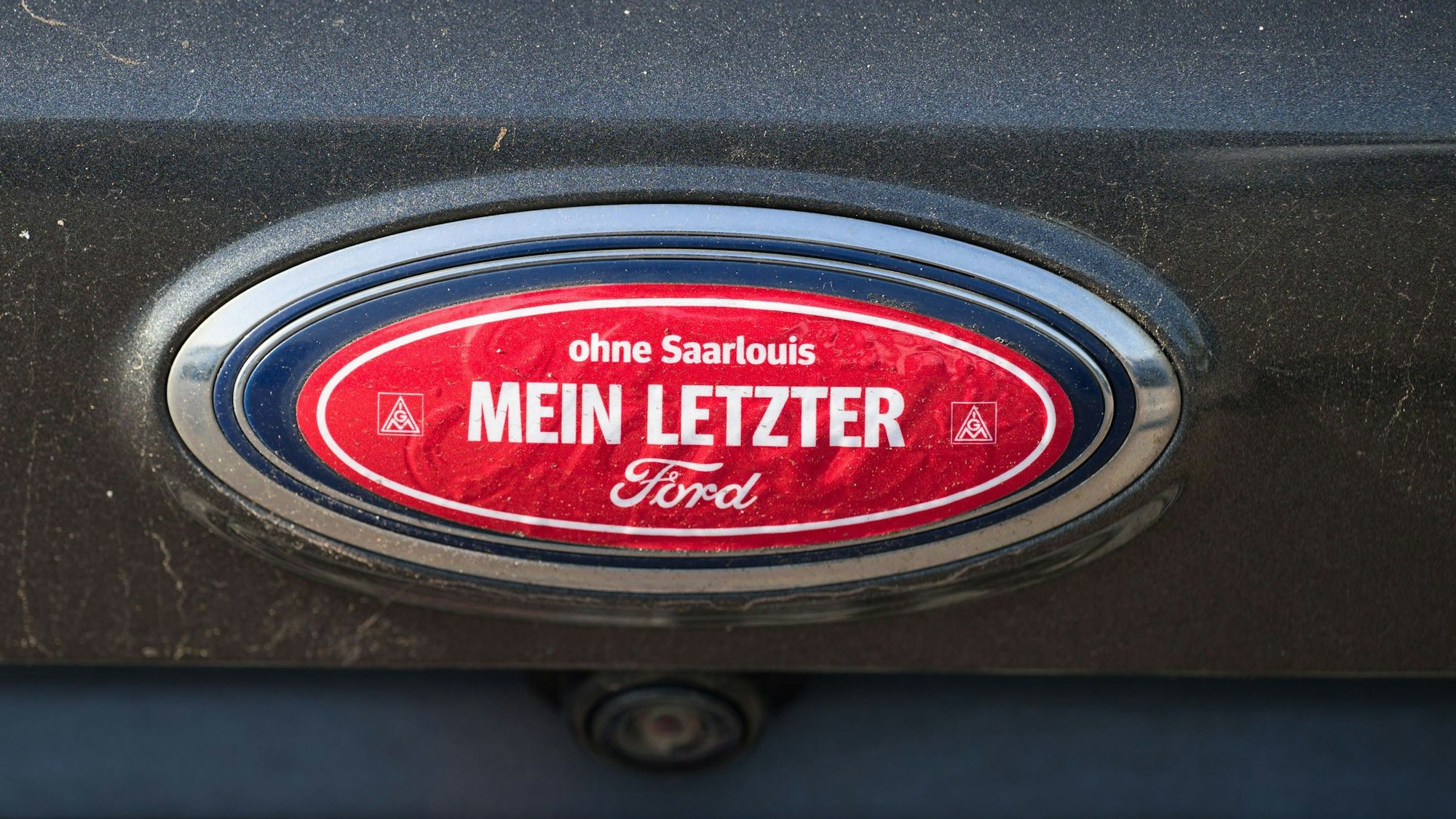 04.10.2023, Saarland, Saarlouis: Ein Aufkleber "ohne Saarlouis mein letzter Ford" klebt auf dem Ford Logo einen Autos, das auf dem dem Parkplatz des Ford Werks in Saarlouis steht. Bei einer Betriebsversammlung hofft die Belegschaft mehr über die Zukunft des Werkes zu erfahren. Foto: Oliver Dietze/dpa +++ dpa-Bildfunk +++