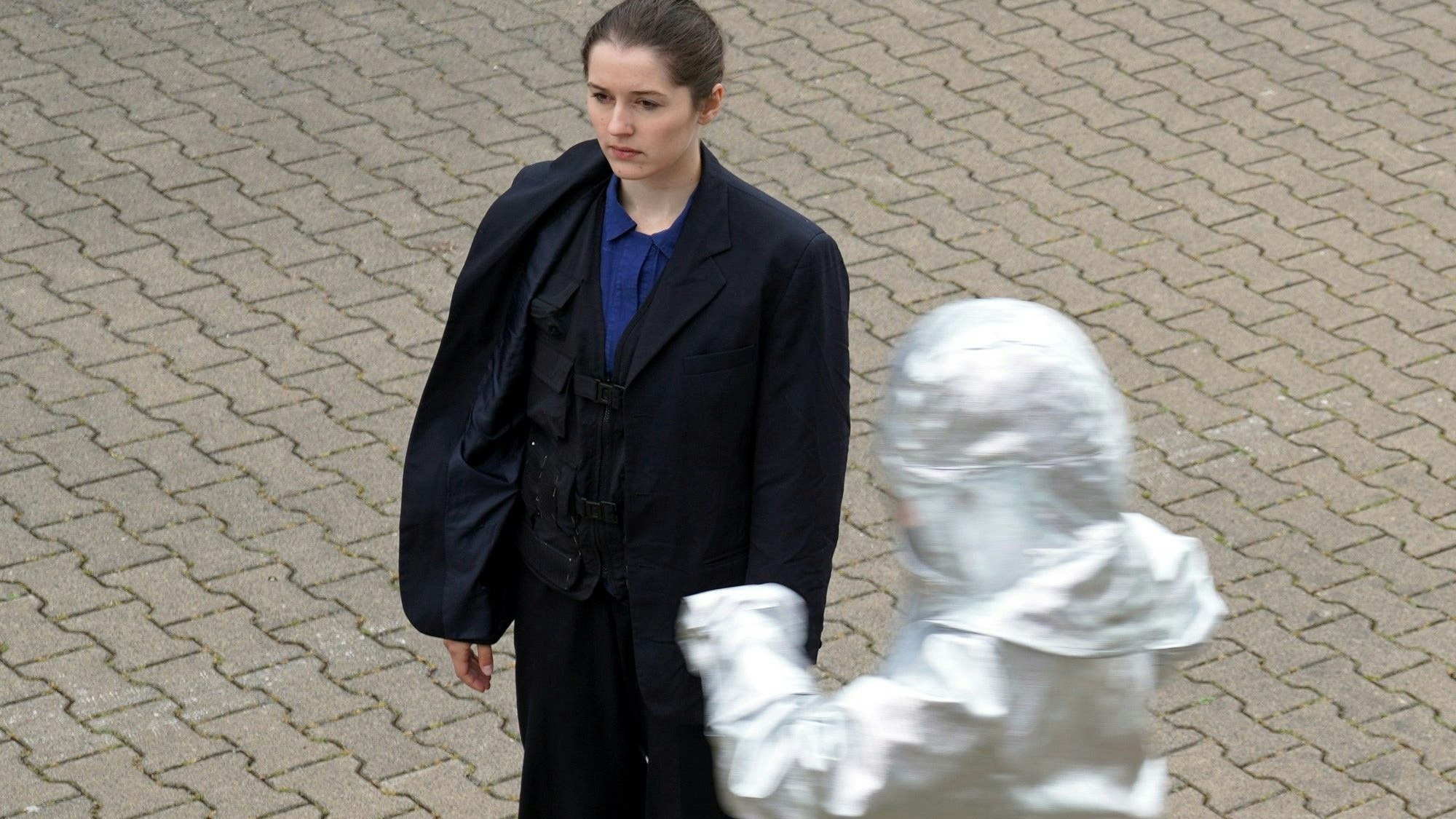 Eine Frau blickt stoisch geradeaus und eine weitere, in einen weißen Schutzanzug gehüllte Person sind auf dem Bild zu sehen.