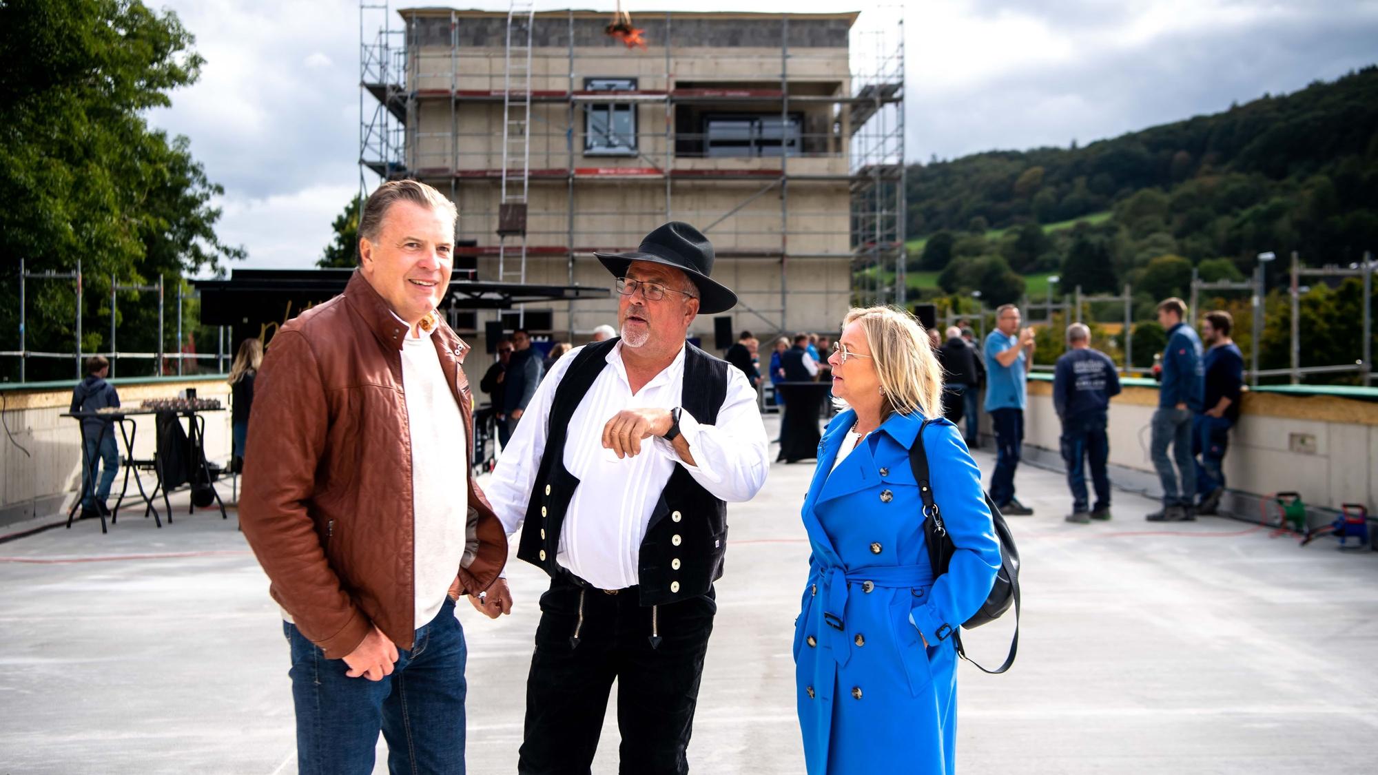 Bauherr Marc Brucherseifer (l.), Zimmermeister Jürgen Dreßen und Bürgermeisterin Sabine Preiser-Marian stehen auf einem betonierten Platz vor einem eingerüsteten und in Bau befindlichen Gebäude.