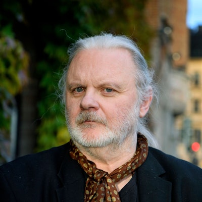 Der norwegische Schriftsteller Jon Fosse posiert für ein Foto in Stockholm. Er hat lange weiße Haare, die er sich hinten zusammengebunden hat, einen Bart, und trägt einen braunen gepunkteten Schal.