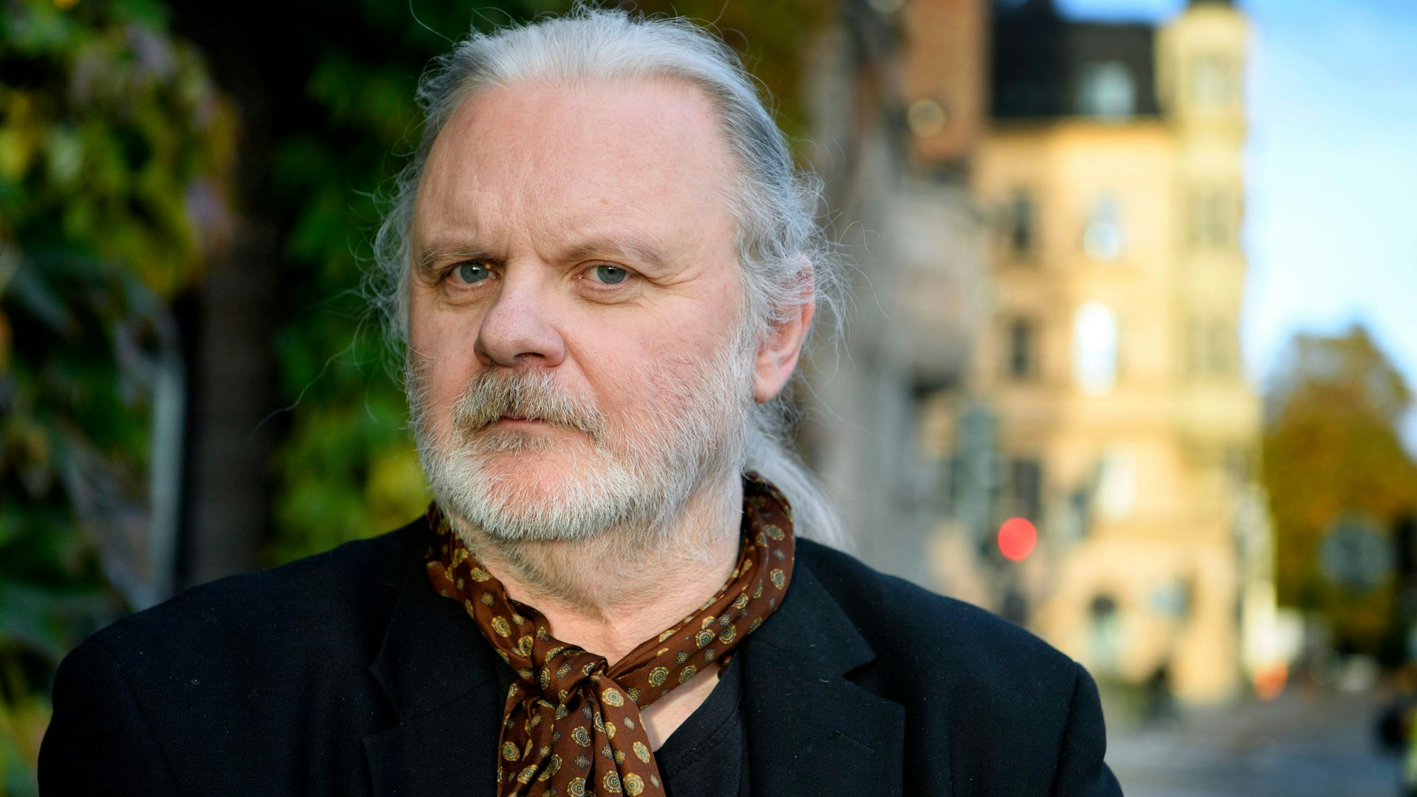 Der norwegische Schriftsteller Jon Fosse posiert für ein Foto in Stockholm. Er hat lange weiße Haare, die er sich hinten zusammengebunden hat, einen Bart, und trägt einen braunen gepunkteten Schal.