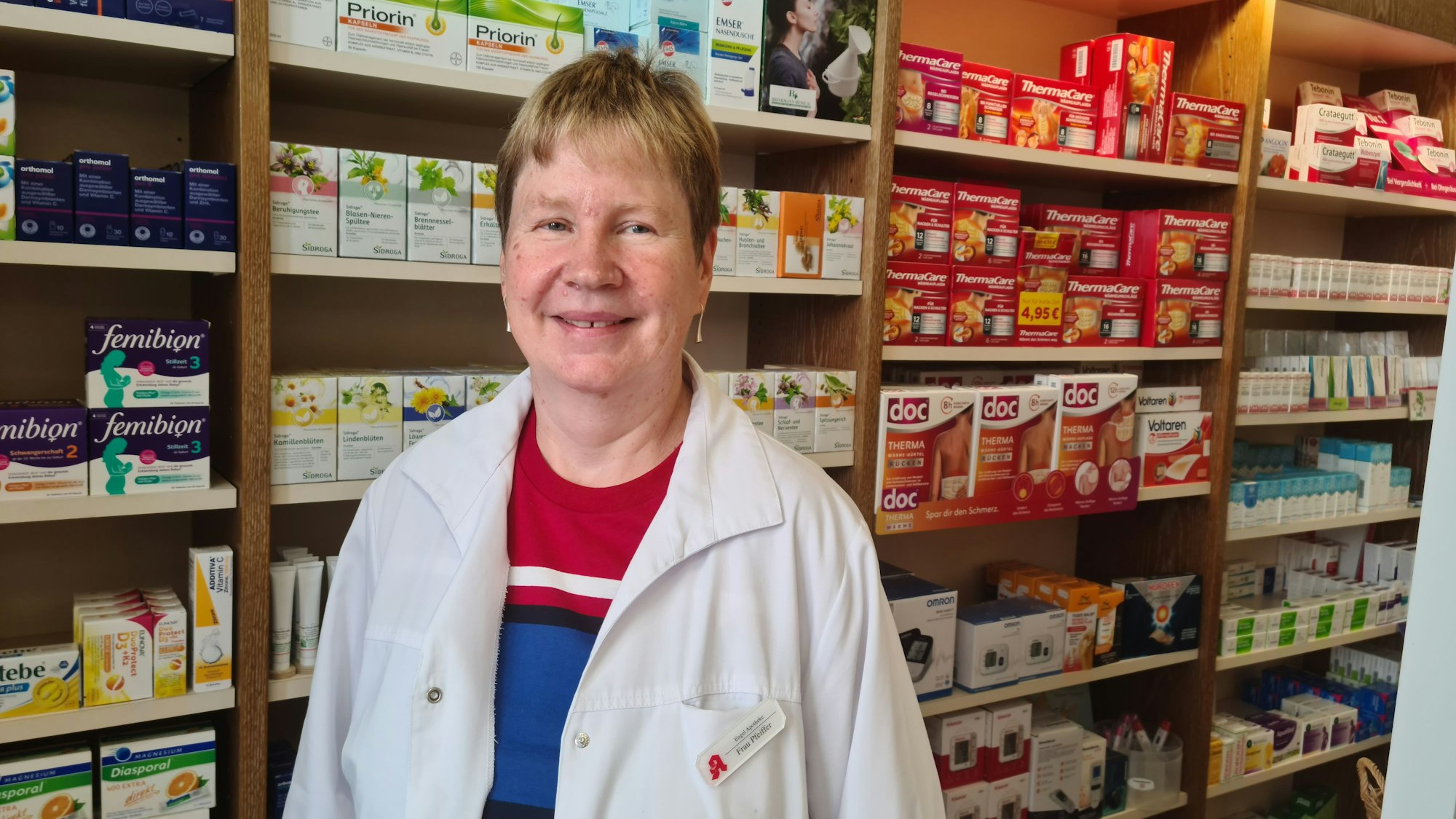 Eine Frau in mittleren Jahren mit kurzem Haar und weißem Kittel über einem gestreiften Pulli steht vor einem Regal mit Arzneimitteln.