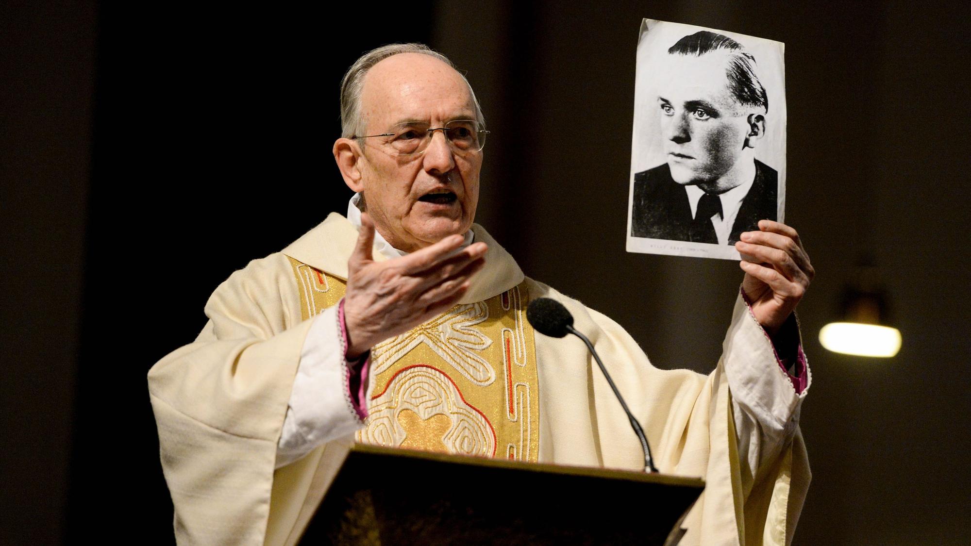 Prälat Helmut Moll steht im Priestergewand an einem Pult und hält eine Fotografie, die Willi Graf zeigt, in der Hand.