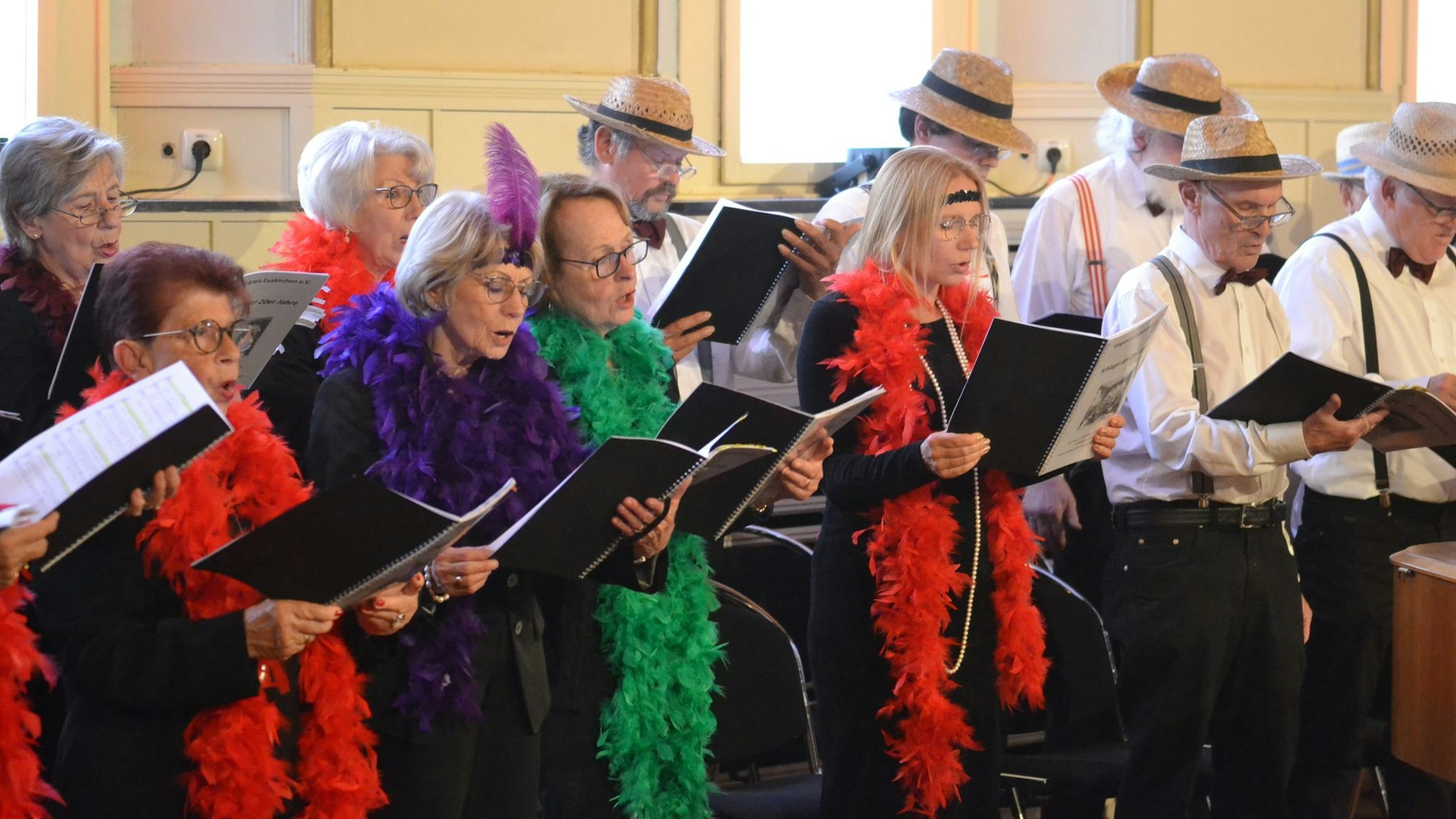 Die Sängerinnen und Sänger des Sängerkreises, die Frauen ausstaffiert mit Federboas, die Männer mit Strohhüten, weißen Hemden und Hosenträgern, bei einem Gesangsvortrag.