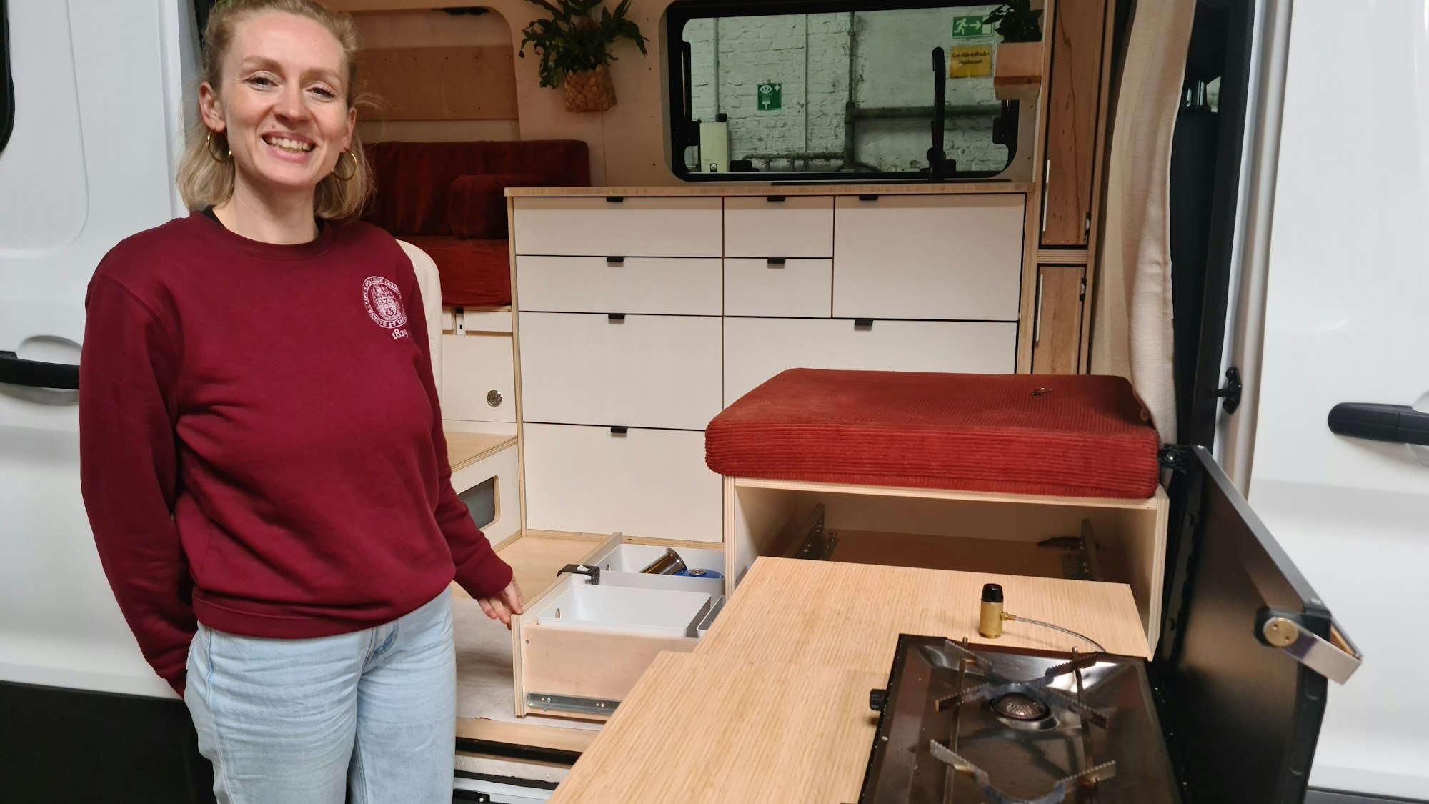 Eine junge Frau steht vor einem Wohnmobil, drinnen ist die Küchenzeile zu sehen, sie steht neben dem Herd, der nach draußen ausgezogen wird.