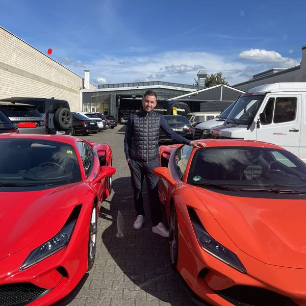 Ein junger Mann mit dunklem Haar steht zwischen zwei roten Ferrari.