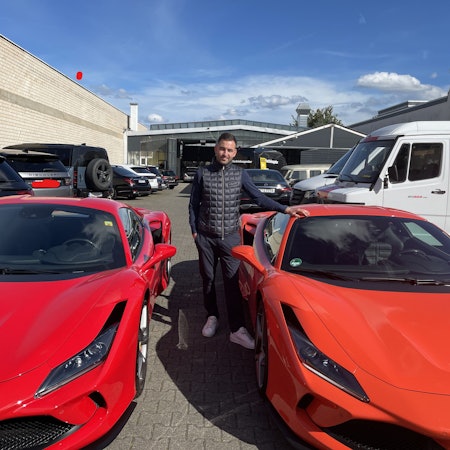 Ein junger Mann mit dunklem Haar steht zwischen zwei roten Ferrari.