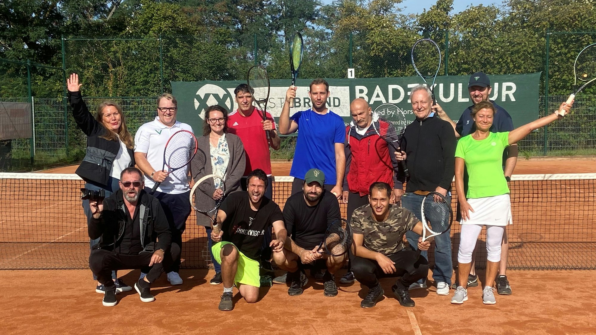 Eine Gruppe Menschen posiert in Sportkleidung und mit Tennisschlägern vor einem Tennisnetz.