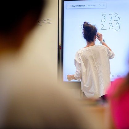Eine Lehrerin schreibt eine Mathematikaufgabe auf eine digitale Schultafel im Klassenraum einer 4. Klasse einer Grundschule.