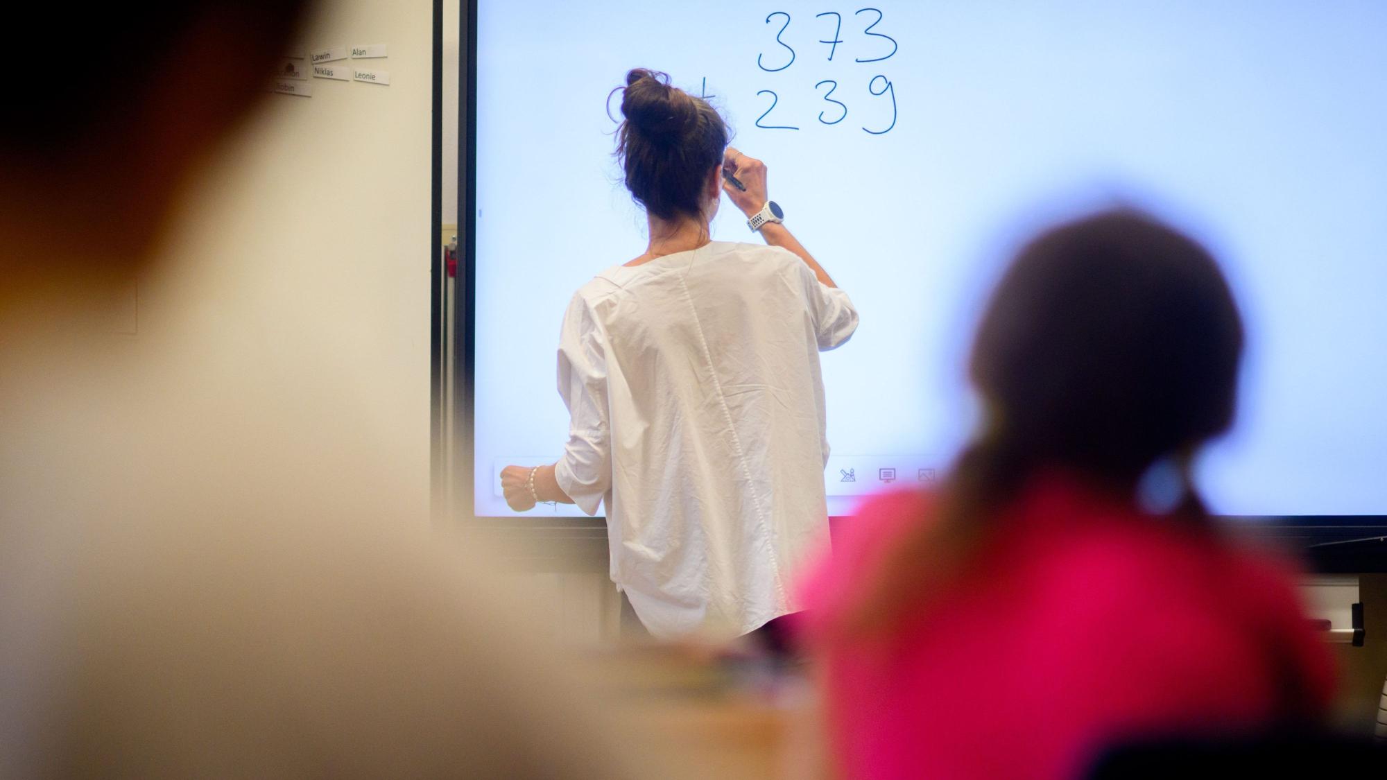 Eine Lehrerin schreibt eine Mathematikaufgabe auf eine digitale Schultafel im Klassenraum einer 4. Klasse einer Grundschule.