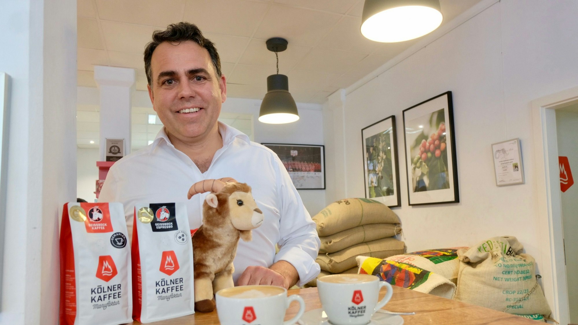Ein Mann schaut in die Kamera. Vor ihm stehen zwei Tüten Kaffee und zwei Kaffeetassen.