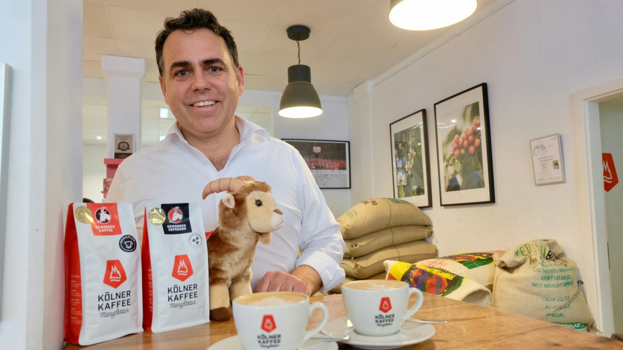Ein Mann mit dunklem Haar steht lachend hinter einem Stehtisch, auf dem zwei Kaffeesorten und ein Plüsch-Geißbock aufgebaut sind, daneben auch zwei Kaffeetassen.