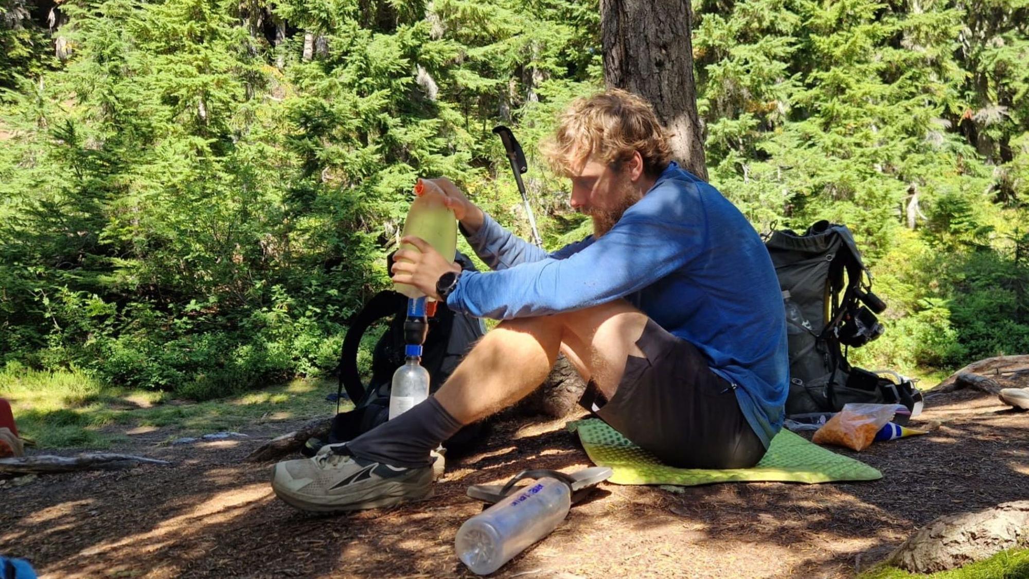 David Flitsch schüttet Wasser in eine Trinkflasche, er sitzt auf dem Boden vor einem Baum.