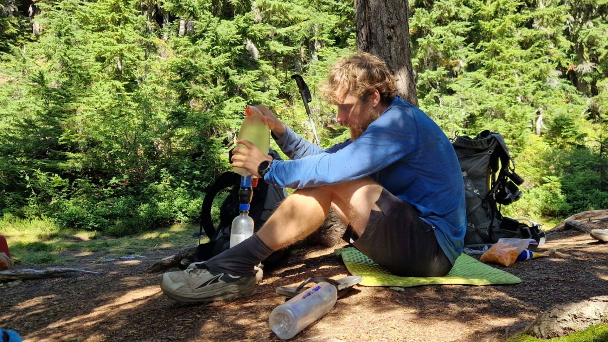 David Flitsch schüttet Wasser in eine Trinkflasche, er sitzt auf dem Boden vor einem Baum.