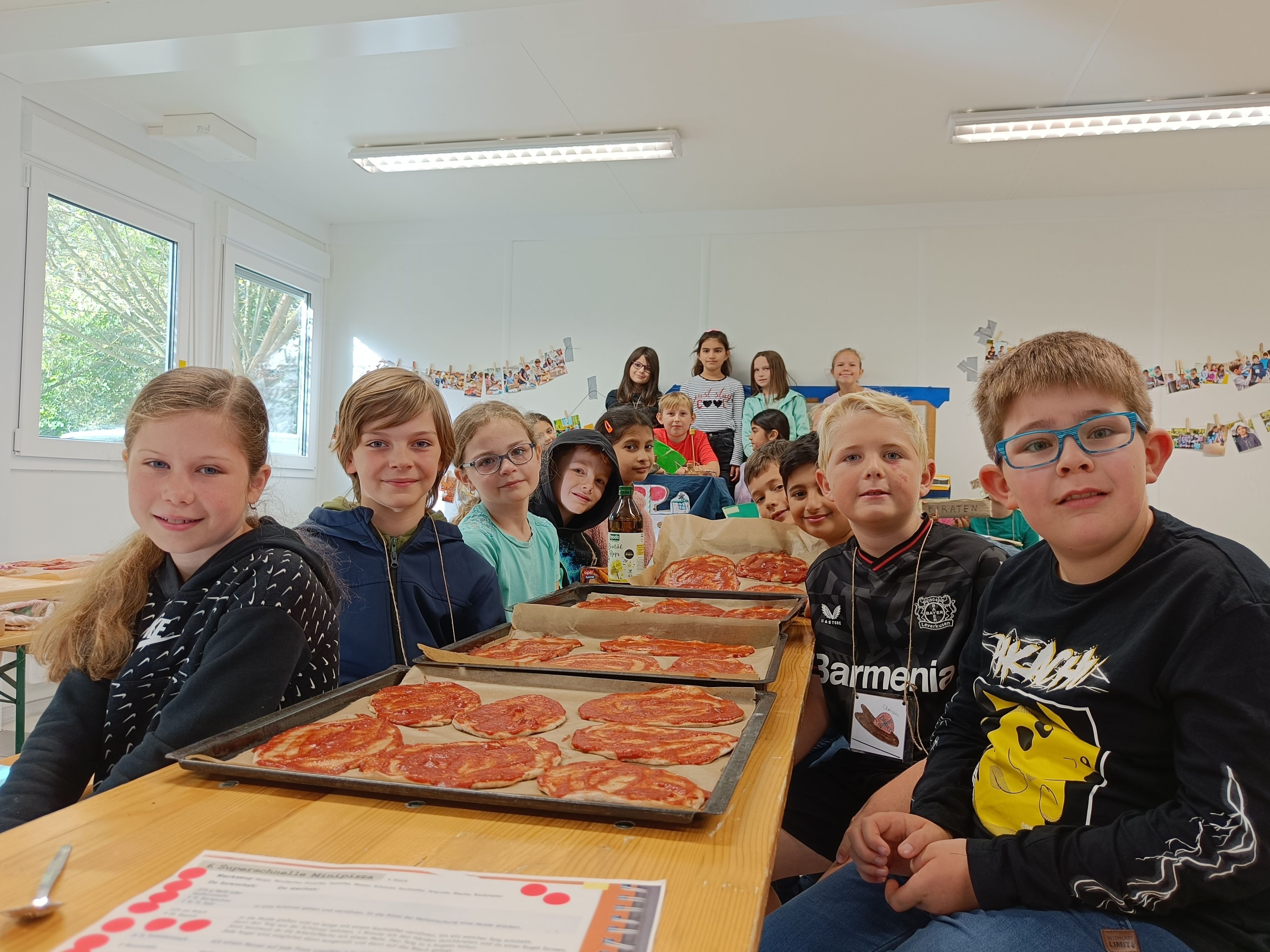 Kinder sitzen an und hinter einem langen Tisch. Vor ihnen stehen Bleche mit noch unbelegter Pizza.