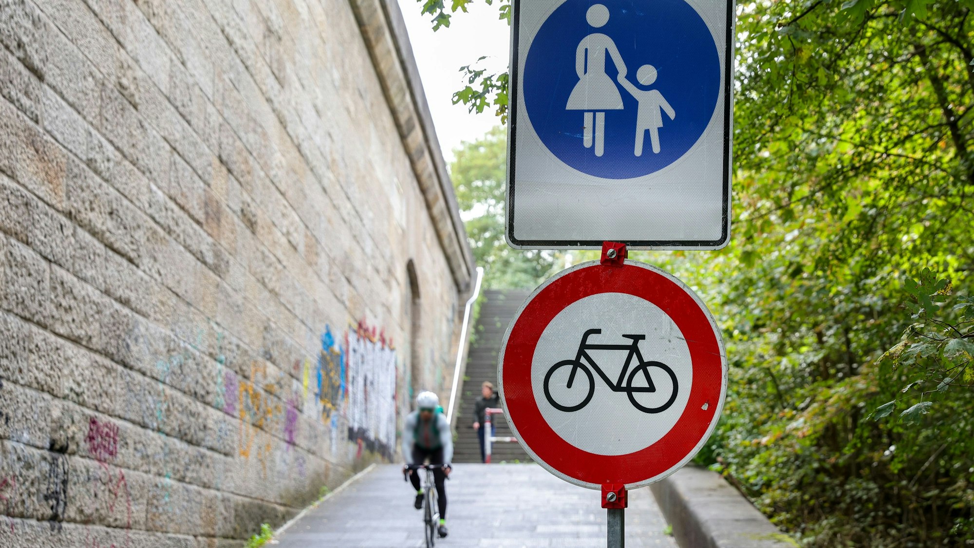 Schilder stehen auf der Auffahrt zur Rodenkirchener Brücke, im Hintergrund ist ein Rennradfahrer zu sehen.