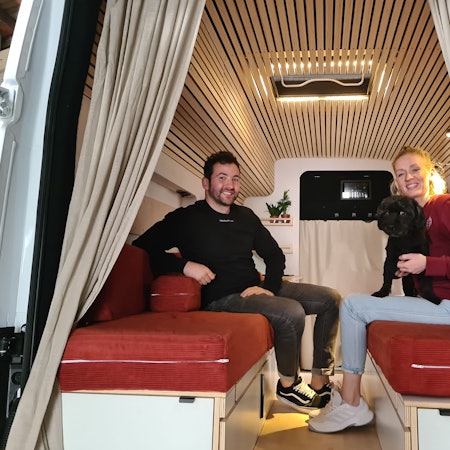 Ein junger Mann mit dunklem Haar und eine junge blonde Frau sitzen sich in einem Transporter auf Polsterbänken gegenüber. Die Frau hat einen schwarzen Hund auf dem Schoß.